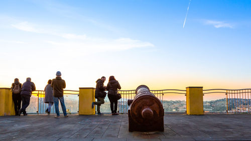 Rear view of people standing on railing by sea against sky