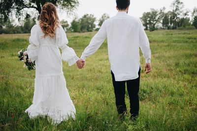 Rear view of wedding couple holding hands while standing on grassland