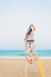Rear view of woman with scarf sitting on lifeguard chair at beach against clear sky