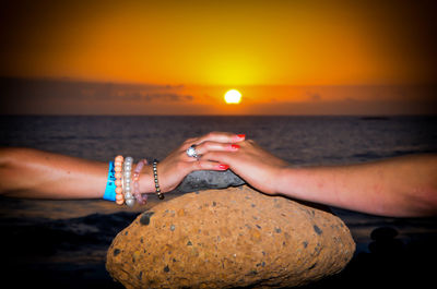 Cropped hand of couple on rock at beach against sky during sunset