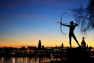 Silhouette person using bow and arrow against illuminated city