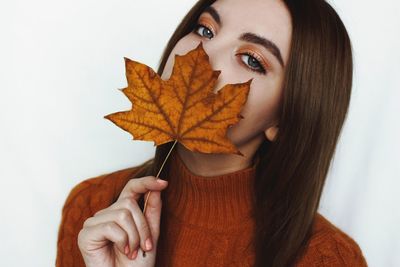 Close-up portrait of young woman holding autumn leaf while standing against white background