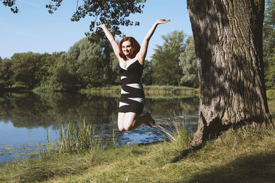 Smiling young woman jumping at lake against trees