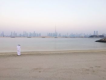 Rear view of man standing on shore by buildings against sky