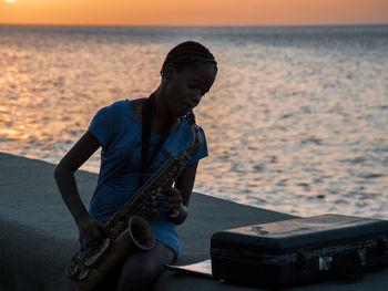 Man playing guitar on sea against sky during sunset
