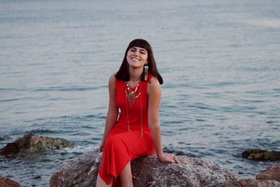 Portrait of smiling beautiful woman wearing red dress while sitting on rock against sea