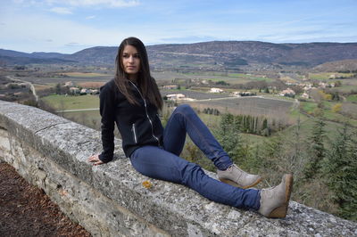 Full length of young woman looking away while sitting on observation point against sky