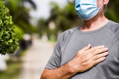 Midsection of senior man wearing mask with hand on chest standing outdoors