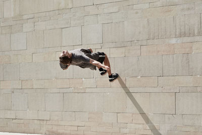 Back view of courageous young male jumping on concrete wall of building while performing dangerous stunt and doing parkour in city