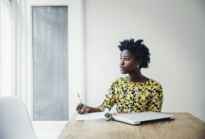 Thoughtful businesswoman writing in book at desk in board room