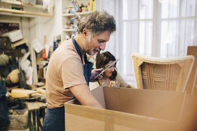 Male craftsperson holding digital tablet while looking in cardboard box at workshop
