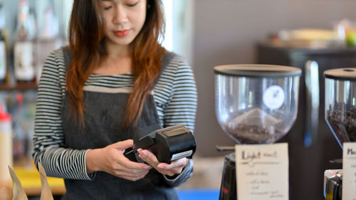 Asian barista using payment terminal, entering drinking cost for customer payment at coffee shop