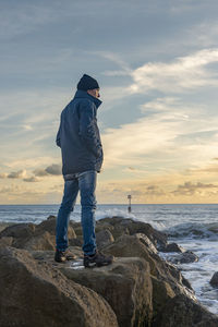 Man standing on rocks looking out to sea, wearing outdoor clothing.