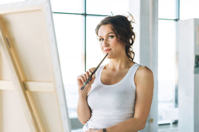 Thinking woman artist in white casual clothes with paint brushes in front of easel in modern gallery