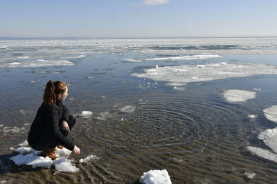 Full length of young woman sitting by frozen lake during winter