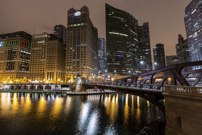 Illuminated bridge over river by buildings against sky at night