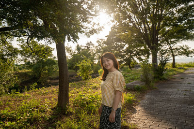 Portrait of woman standing on footpath against tree