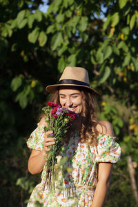 Smiling woman holding flowers while standing against trees