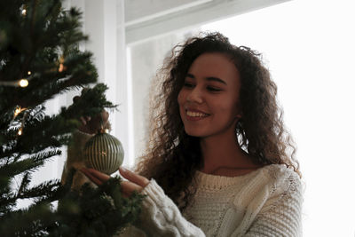 Happy young woman decorating christmas tree with bauble at home