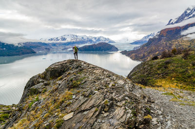 Mid distance view of hiker standing on cliff by lake against cloudy sky