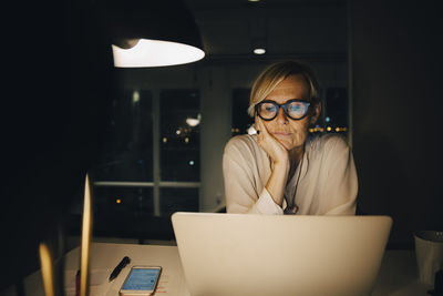 Tired mature female professional staring at laptop while sitting with hand on chin working late in office