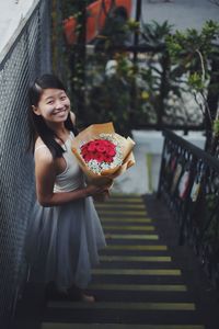Portrait of smiling young woman holding bouquet while standing on stairs