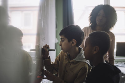 Boys doing mathematics on white board while teacher examining in classroom