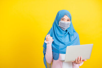 Young woman wearing mask holding laptop standing against yellow background