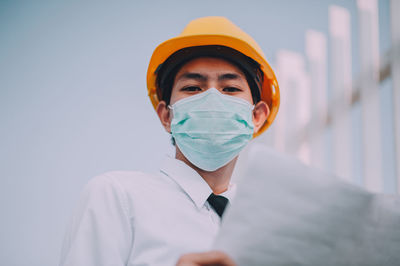 Low angle portrait of male architect wearing mask and helmet against sky