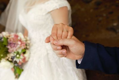 Midsection of bride and groom holding hands with wedding ring holding bouquet