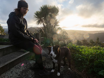 Full length of woman sitting on fence by dog against sky