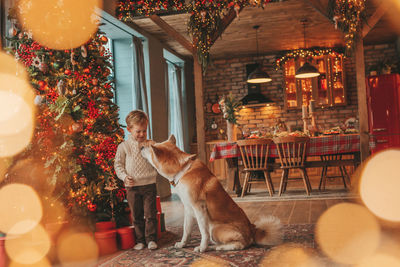 Candid authentic happy little boy in knitted beige sweater hugs dog with bow tie at home on xmas