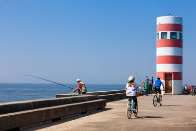 People riding bicycle on lighthouse by sea against clear sky