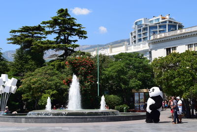 People standing by fountain on street against trees