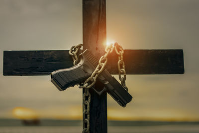 Close-up of handgun tied with chain on cross against sky during sunset