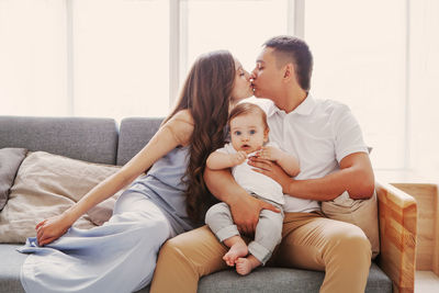 Mom and dad kissing while the child looking at camera. happy family sitting home on couch