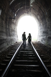 Rear view of female friends holding hands while standing on railroad track in tunnel