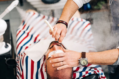 From above of crop anonymous beauty master in wristwatch shaving beard of client with straight razor during steam vapor treatment in hairdressing salon