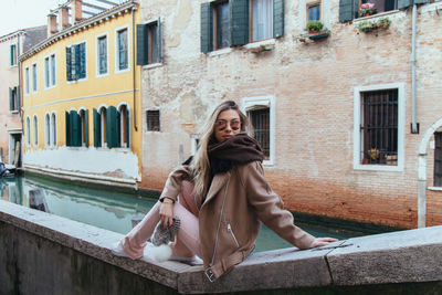 Side view of young woman sitting on retaining wall against building