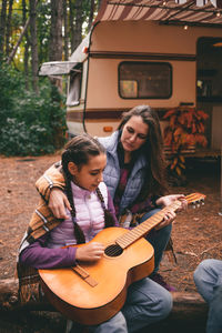 Mom and daughter near the camping trailer in the forest play the guitar