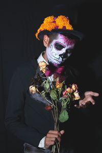 Man with halloween make-up gesturing while holding dry flowers against black background