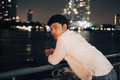 Man leaning on railing by river in illuminated city at night