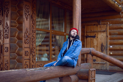 Portrait of a village man with a beard in a red hat stands at a wooden house in autumn