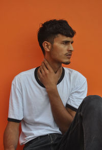 A handsome indian young guy looking away while sitting outside against orange wall with copy space
