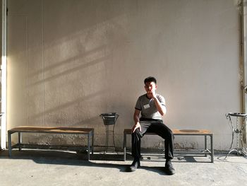 Full length of young man sitting on bench against wall