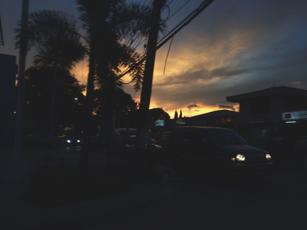 sunset, silhouette, transportation, sky, building exterior, car, built structure, land vehicle, street, architecture, mode of transport, power line, road, electricity pylon, city, dusk, tree, street light, orange color, dark