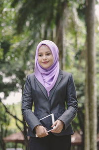 Young woman in hijab with digital tablet standing against trees