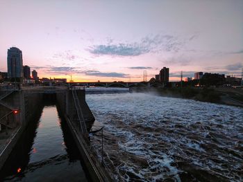 Bridge over river by buildings against sky during sunset