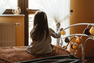 Child sitting on the bed at home. dreamy sunny mood. girl looking though the window from the back