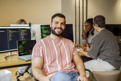 Portrait of smiling male coding professional sitting near desk with colleagues at office startup